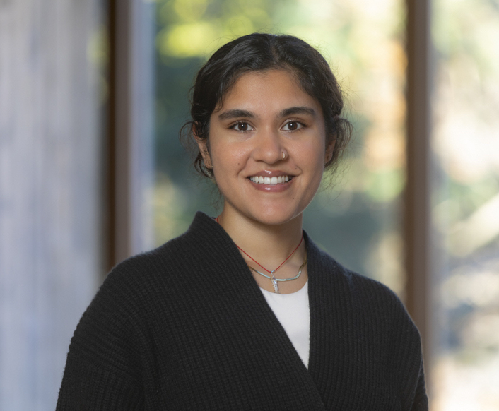 Professional headshot. Vinitha, an Indian woman, smiles at the camera. She has black curly hair that is tied back and is wearing a black cardigan.